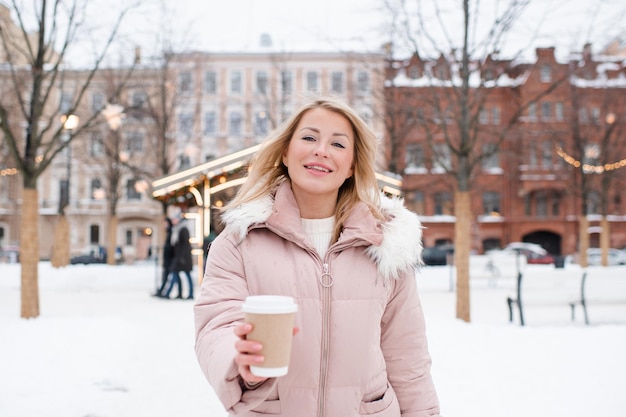 Junge schöne lächelnde blonde Frau mit Mantel, die über weihnachtlichen Stadtdekorationen und Glühlichtern steht, hält eine Tasse Kaffee im Freien Wintergruß Ferienzeit
