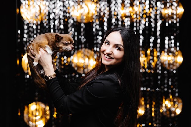 Junge schöne lächelnde brünette Frau in schwarzer Jacke, die ihren kleinen Hund in ihren Händen hält Sorglose Frau posiert in der Nähe einer glänzenden Lametta-Wand im Studio mit Hund