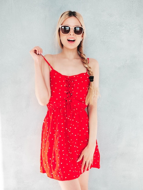 Junge schöne lächelnde Frau im trendigen roten Sommerkleid Sexy sorglose blonde Frau posiert in der Nähe der Wand im Studio Positives Modell, das Spaß hat Fröhlich und glücklich Am sonnigen Tag