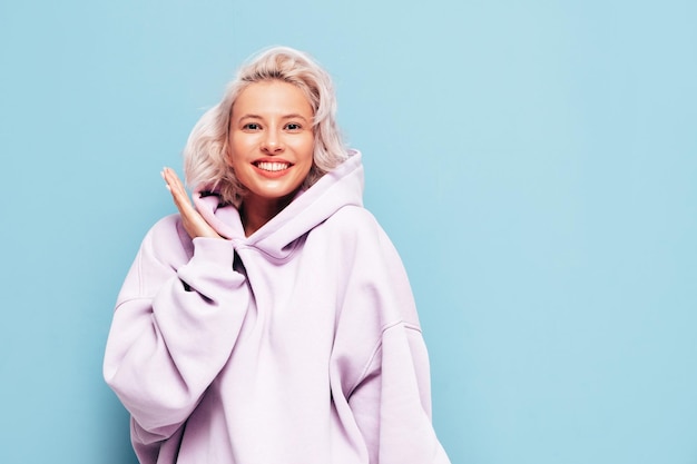 Junge schöne lächelnde Frau im trendigen Sommer-Hoodie Sexy sorglose Frau posiert in der Nähe der blauen Wand im Studio Positives blondes Modell, das Spaß hat und verrückt wird Fröhlich und glücklich