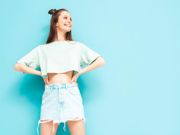 Junge schöne lächelnde Frau im trendigen Sommerjeansrock sorglose Frau posiert in der Nähe der blauen Wand im Studio Positives Modell, das sich drinnen amüsiert Fröhlich und glücklich