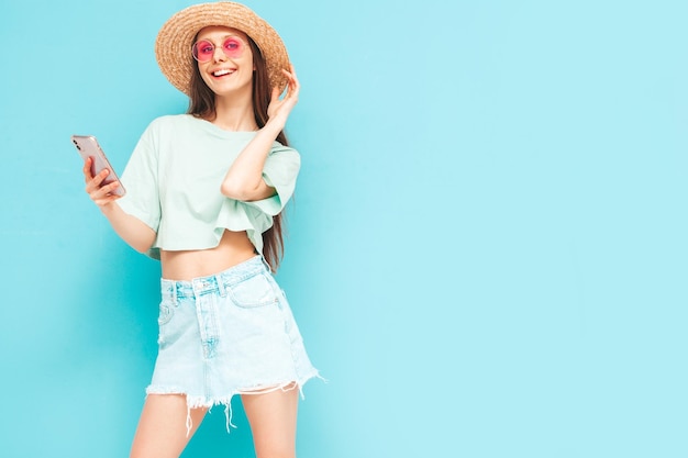 Junge schöne lächelnde Frau im trendigen Sommerrock Sexy Frau mit Posing in der Nähe der gelben Wand im Studio Positives Modell, das Spaß hat In Sonnenbrille und Hut Blick auf Smartphone mit Apps