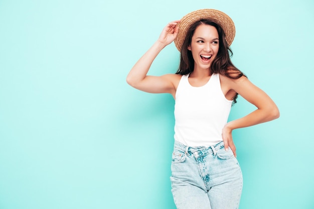 Junge schöne lächelnde Frau in trendiger Sommerkleidung Sexy sorglose Frau posiert in der Nähe der blauen Wand im Studio Positives brünettes Modell, das Spaß hat Fröhlich und glücklich In Hut