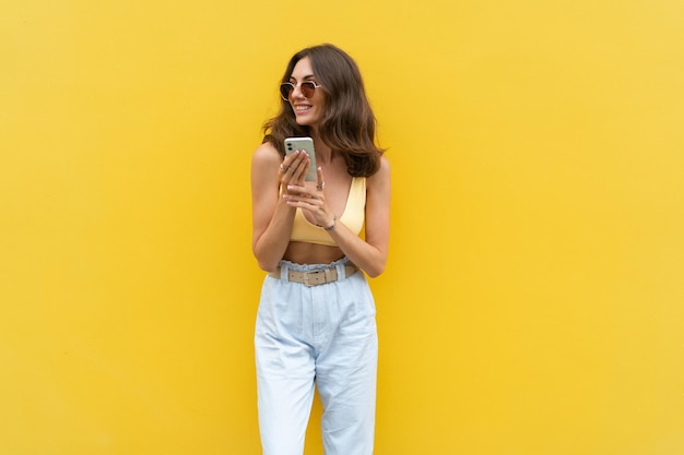 Junge schöne lächelnde Frau mit Handy Sorglose Frau, die auf der Straße in der Nähe der gelben Wand posiert Positives Modell im Freien mit Sonnenbrille Glücklich und aufgeregt