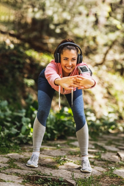 Junge, schöne, lächelnde Läuferin, die Musik hört, während sie sich vor dem Joggen in der Natur dehnt.