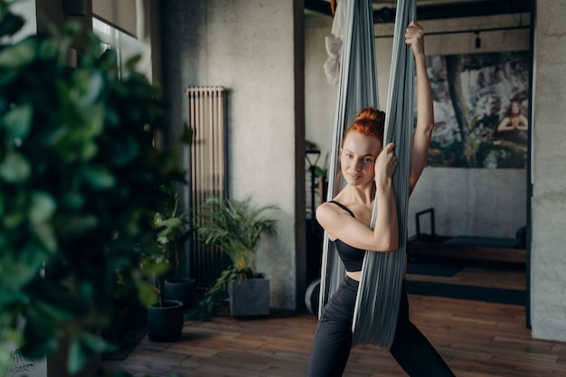 Junge schöne lächelnde rothaarige Frau in aktiver Kleidung, die sich nach dem Üben von Fliegenyoga im Fitnessstudio ausruht, sich auf grauer Hängematte entspannt und in die Kamera schaut Gesundheits-, Sport- und Yogakonzept
