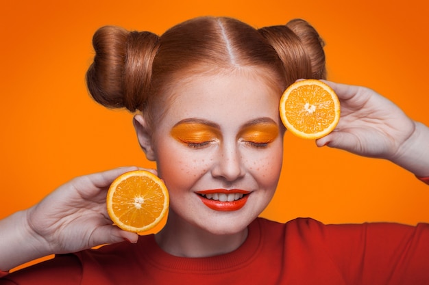 Junge schöne lustige Mode-Modell mit Orangenscheibe auf orangem Hintergrund. mit orangem Make-up und Frisur und Sommersprossen. Studioaufnahme, Blick in die Kamera mit geschlossenen Augen und lächelnd.