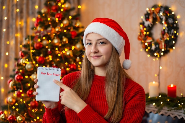 Junge schöne Mädchen in roter Weihnachtsmütze mit Notizen, die mit dem Finger auf die Inschrift Neujahrsziele zeigen Konzept der Wunschliste oder To-do-Liste für das kommende Jahr