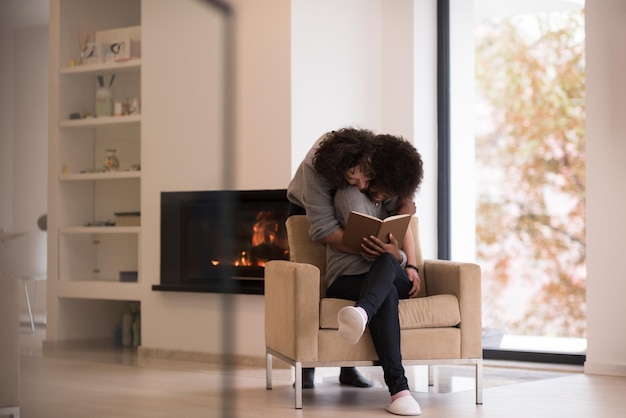 Junge schöne multiethnische Paare, die sich zu Hause vor dem Kamin umarmen, wenn sie am Herbsttag ein Buch lesen