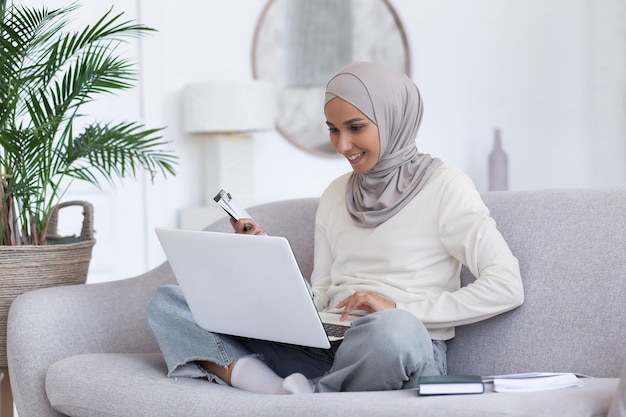 Junge schöne muslimische Frau im grauen Hijab, die zu Hause auf dem Sofa sitzt, Studentin mit