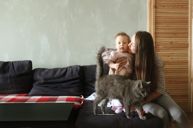 Junge schöne Mutter, die auf einer Couch zu Hause sitzt und ihr süßes Baby umarmt und küsst