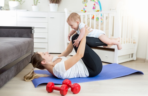 Junge schöne Mutter macht Yoga-Übungen mit ihrem Baby auf dem Boden im Wohnzimmer