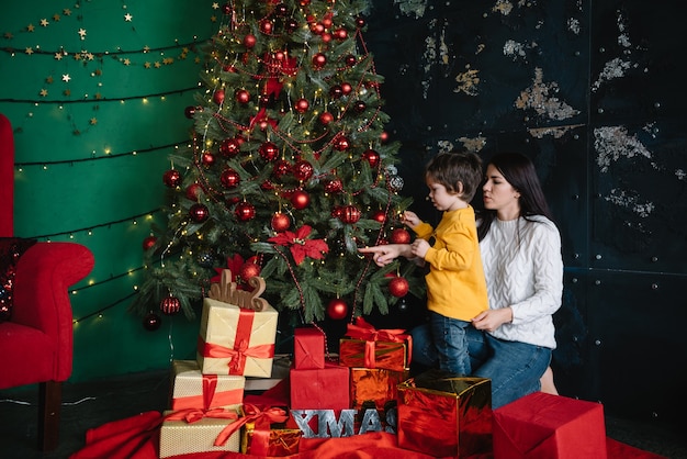 Junge schöne Mutter mit dem niedlichen Sohn nahe Weihnachtsbaum mit Geschenken. Fröhliche Weihnachten