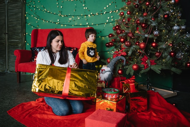 Junge schöne Mutter mit dem niedlichen Sohn nahe Weihnachtsbaum mit Geschenken. Fröhliche Weihnachten