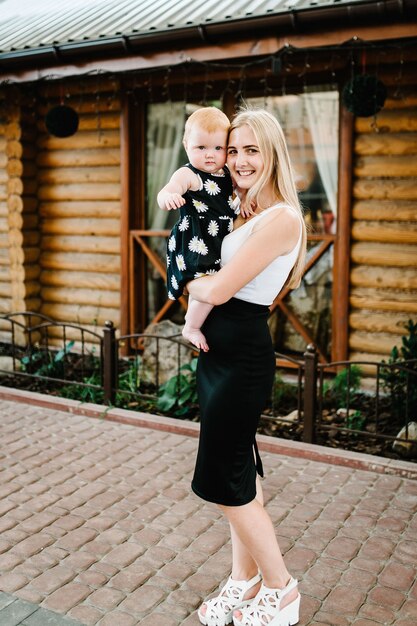 Junge schöne Mutter mit einem kleinen Mädchen auf ihren Händen, die über einem Holzhaus stehen. Baby.