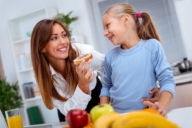 Junge schöne Mutter und ihre süße Tochter beim Frühstück in der Küche. Mutter füttert Tochter mit Brotscheibe mit Pastete.