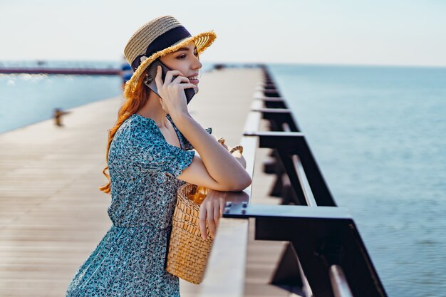 Junge schöne rothaarige Mädchen sprechen auf dem Smartphone, während sie auf dem Pier stehen und in Mode tragen...