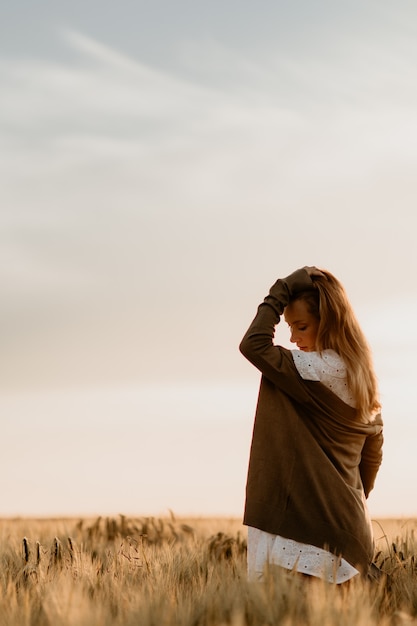 Junge schöne schwangere Frau im weißen Kleid und in der Jacke, die nach unten schaut und ihre Haare im Feld an einem sonnigen Sommertag berührt. Natur auf dem Land. Wunder Erwartung. Sonnenuntergang auf Isolation.