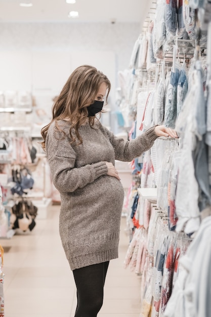 Junge schöne schwangere Frau in der Hygienemaske, die Babykleidung für Neugeborene wählt.