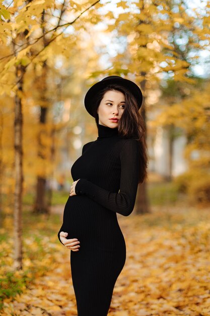 Junge schöne schwangere Frau in einem Hut mit dunklen Haaren in einem schwarzen engen Kleid posiert