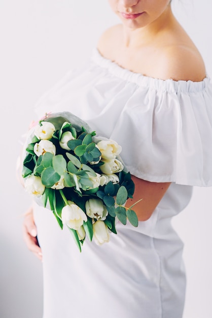 Junge schöne schwangere Frau in einem weißen Kleid mit einem Blumenstrauß der weißen Tulpen