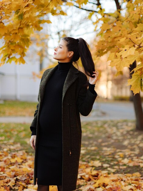 Junge schöne schwangere Frau mit dunklen Haaren in einem schwarzen engen Kleid posiert auf einem Herbst