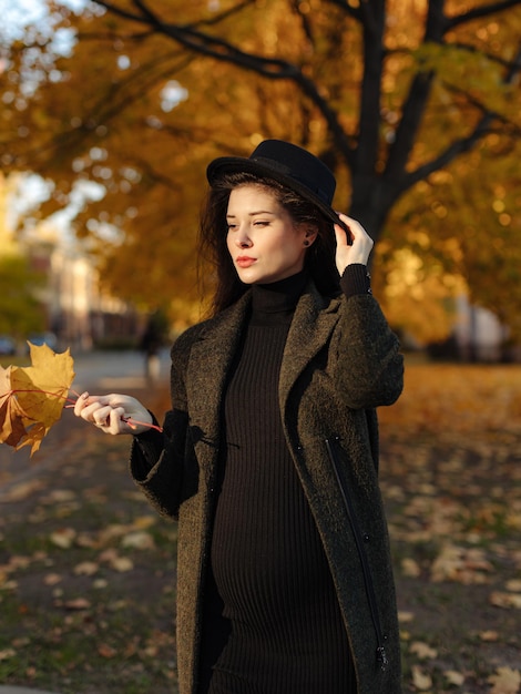 Junge schöne schwangere Frau mit Hut mit dunklen Haaren in einem schwarzen engen Kleid und Mantel posiert