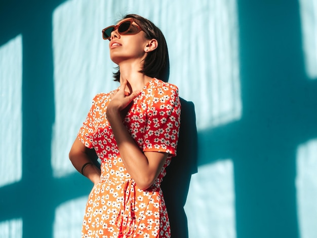 Junge schöne selbstbewusste Frau im trendigen roten Sommerkleid Sexy sorglose Frau posiert in der Nähe der blauen Wand im Studio Positives Modell, das Spaß hat Fröhlich und glücklich Am sonnigen Tag Schatten vom Fenster