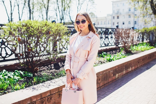 Junge schöne stilvolle Frau, die im weißen Kleid und im rosa Mantel in der Stadt geht und aufwirft. Sommerporträt im Freien der jungen noblen Frau in der Sonnenbrille