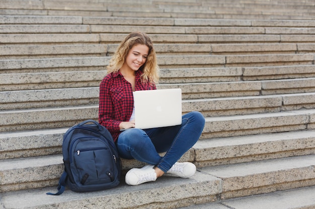 Junge schöne Studentin, die draußen auf der Treppe sitzt und mit Laptop arbeitet und sich auf Prüfungen auf dem Universitätscampus vorbereitet. Technologie, Bildung und Fernarbeitskonzept, Kopierraum