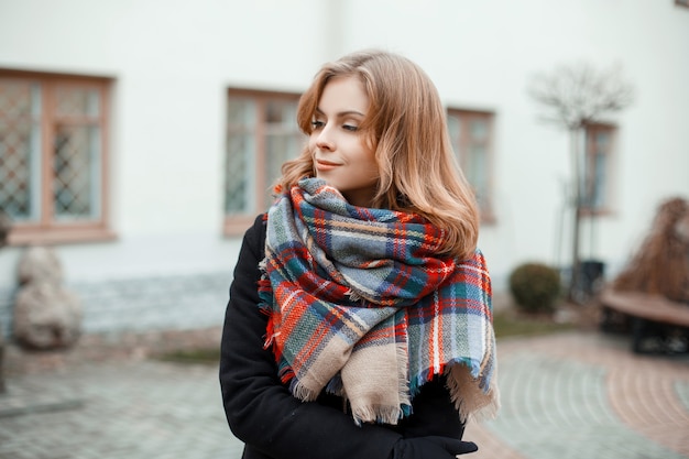 Junge schöne süße Frau in einem trendigen warmen Wintermantel mit einem stilvollen karierten Wollschal steht an einem warmen Herbsttag