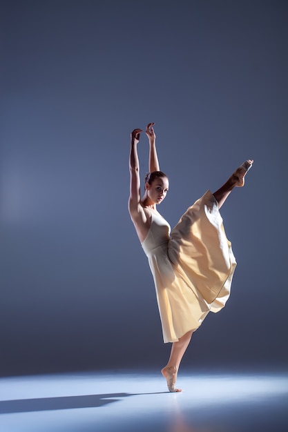 Junge schöne Tänzerin im beigefarbenen Kleid tanzt auf grauem Studiohintergrund