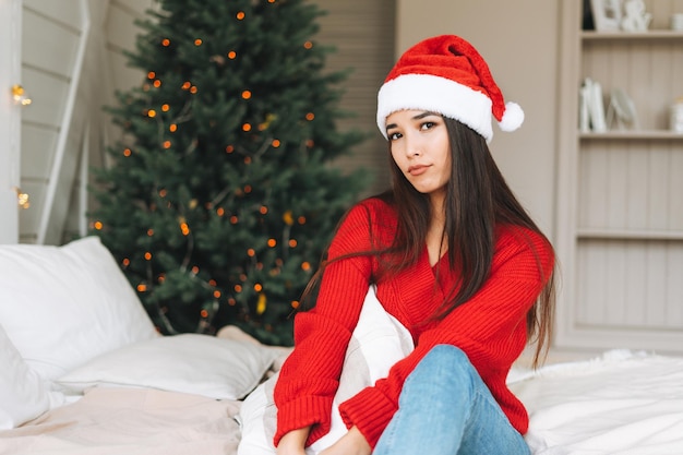 Junge schöne traurige unglückliche asiatin mit dunklen langen haaren in kuscheligem rotem strickpullover und weihnachtsmütze sitzt auf dem bett im zimmer mit weihnachtsbaum zu hause