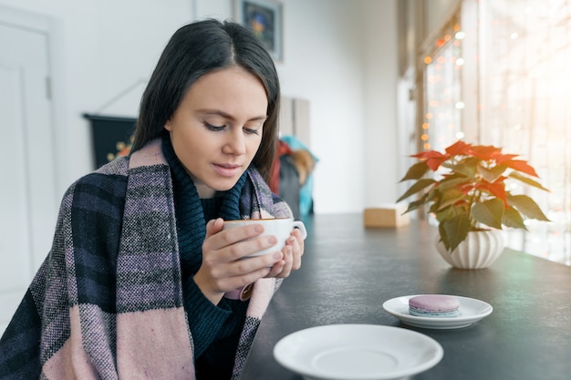 Junge Schönheit in der Wintersaison, die mit dem heißen Kaffee und warmer Decke sitzen in der Kaffeestube nahe dem Fenster aufwärmt.