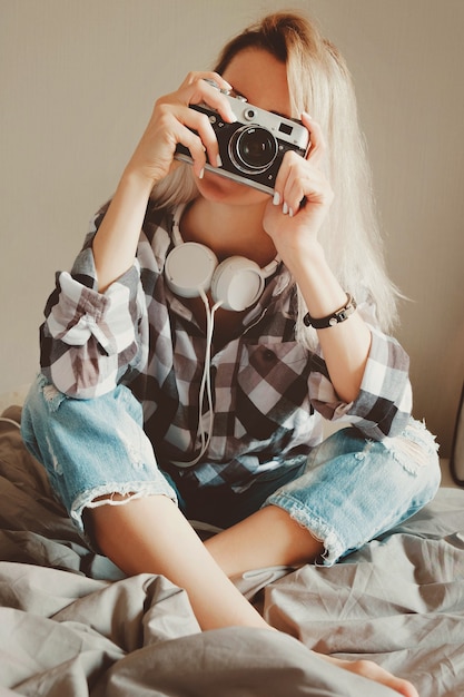 Junge Schönheitsfrau im Freizeithemd, die ein Foto mit ihrer Weinlesekamera auf einem Bett sitzend macht