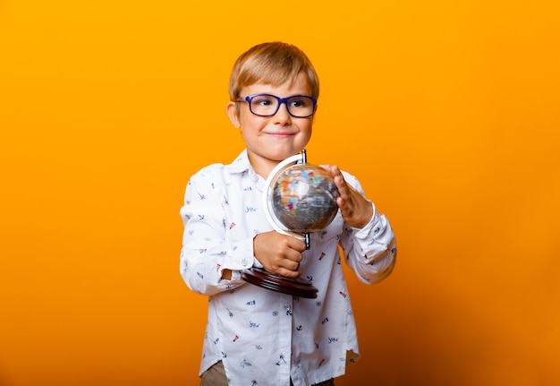 Junge Schuljunge 7 Jahre alter Schuljunge in Gläsern mit einem Globus in den Händen lächelnd auf einem gelben Hintergrund