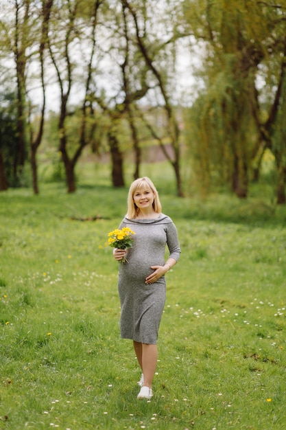 Junge schwangere blonde frau im grauen kleid, schwangeres mädchen im frühjahr auf einem spaziergang, mutterschaftskonzept