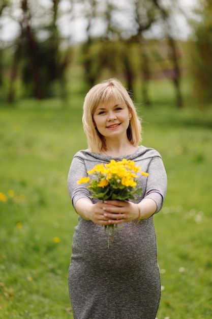 Junge schwangere blonde Frau im grauen Kleid, schwangeres Mädchen im Frühjahr auf einem Spaziergang, Mutterschaftskonzept