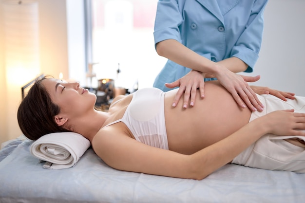 Junge schwangere Frau, die Bauchmassage im Spa-Center erhält. Patientin wird von einem professionellen Therapeuten mit schönen Händen behandelt. Körper positiv, Harmonie, gesunder Lebensstil. Seitenansicht