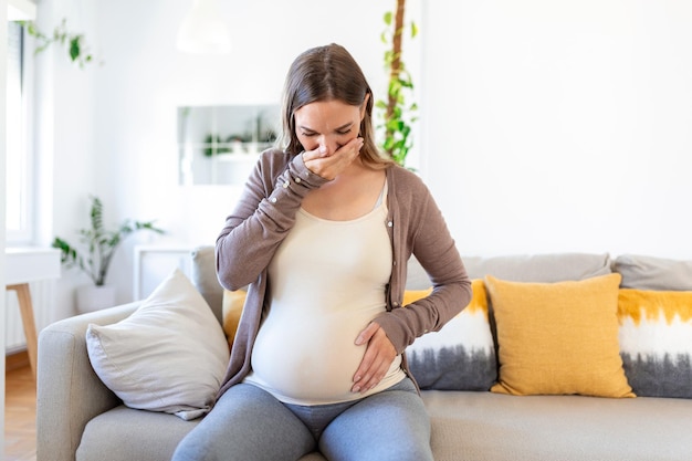 Junge schwangere Frau, die zu Hause an Toxikose leidet Mutterschaftsdame, die morgendliche Übelkeit hat, die den Mund mit den Händen bedeckt, während sie auf der Couch im Wohnzimmer sitzt Mutter erwartet ein ungeborenes Baby