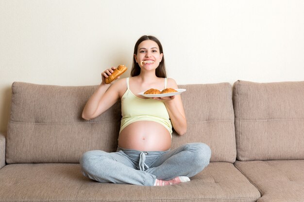 Junge schwangere Frau genießt es, Croissant auf dem Sofa zu essen