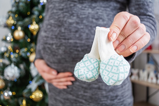 Foto junge schwangere frau mit geschwollenem bauch und babysocken in ihrer hand, die gegen weihnachtsbaum aufwirft