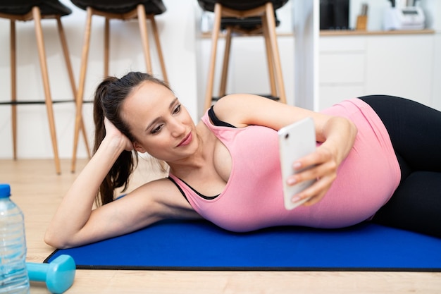 Junge schwangere Frau mit Handy während des Trainings im Wohnzimmer Kaukasische Frau beim Üben