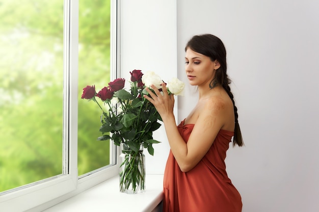 Junge schwangere Frau steht am Fenster neben einem Strauß Rosen in einer Vase
