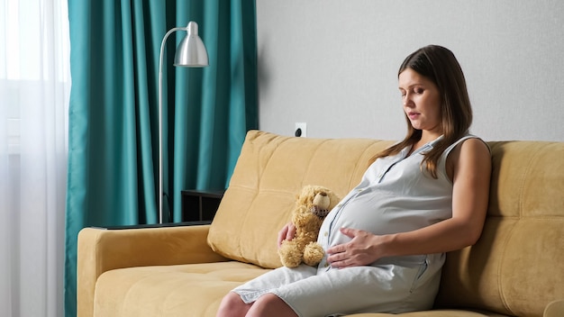 Junge schwangere Frau streichelte den Bauch mit Spielzeugbären beim Sitzen auf dem Sofa.