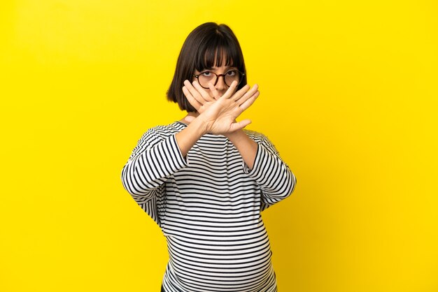 Junge schwangere frau über isoliertem gelbem hintergrund, die mit der hand eine stoppgeste macht, um eine handlung zu stoppen