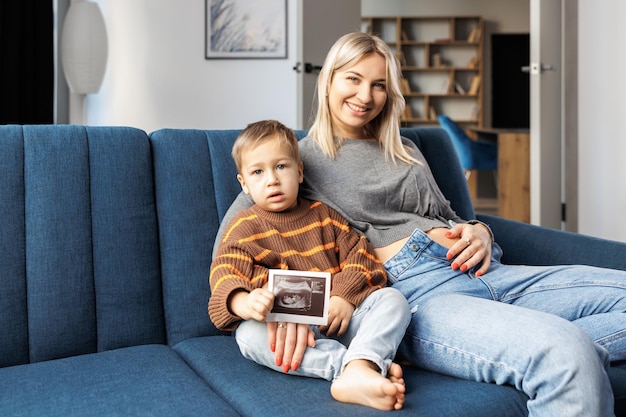 Junge schwangere Frau und ihr Sohn sitzen zu Hause auf dem Sofa und Junge hält Ultraschalluntersuchung in der Hand. Sie verbringen Zeit miteinander und genießen die Kommunikation