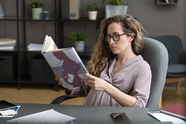 Junge schwangere Geschäftsfrau liest Buch über Schwangerschaft und Geburt