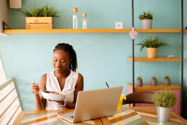 Junge schwarze Frau, die ein gesundes Frühstück hat, während sie im Café am Laptop arbeitet