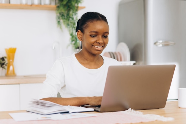 Junge schwarze Frau, die in der Küche sitzt und von zu Hause aus am Laptop arbeitet